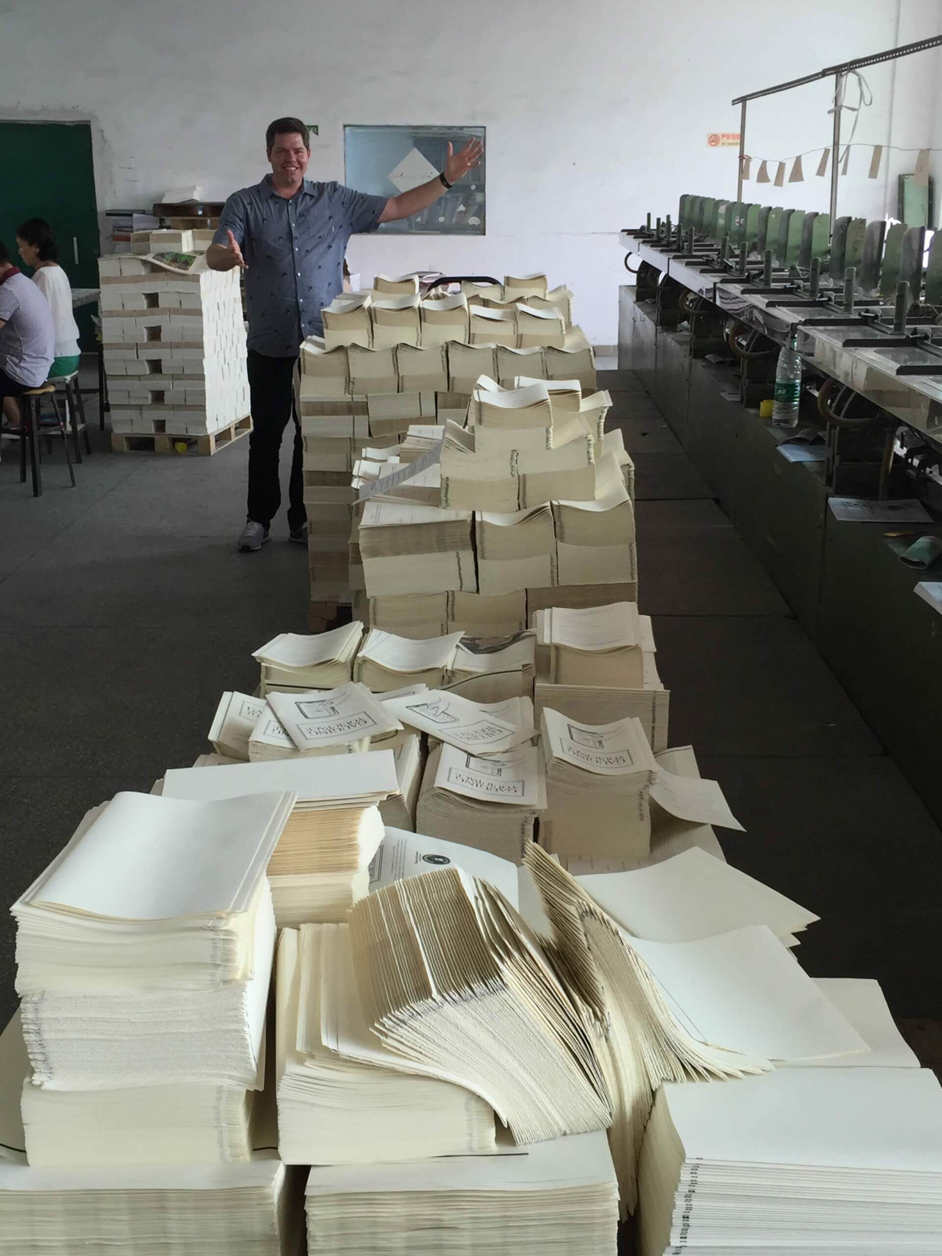 A picture of Richie in a factory, standing behind a stack of hundreds (if not thousands) of journals before they receive their covers. The stack extends to waist height and is at least twelve feet long.