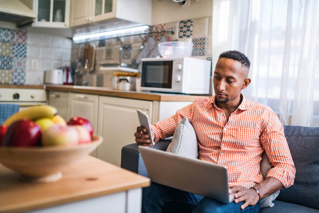 man sitting on couch near a sunny window next to kitchen looking at a laptop and smartphone, with a fruit bowl in the nearground